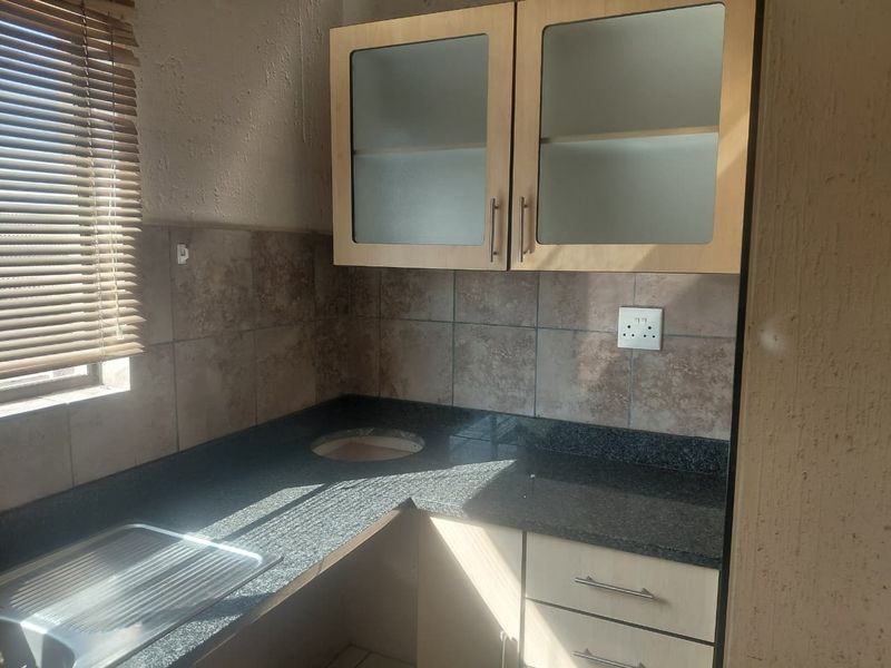 A compact kitchen area featuring granite countertops, stainless steel sink, tiled backsplash, and light wood-finish cabinetry with glass-fronted upper units.