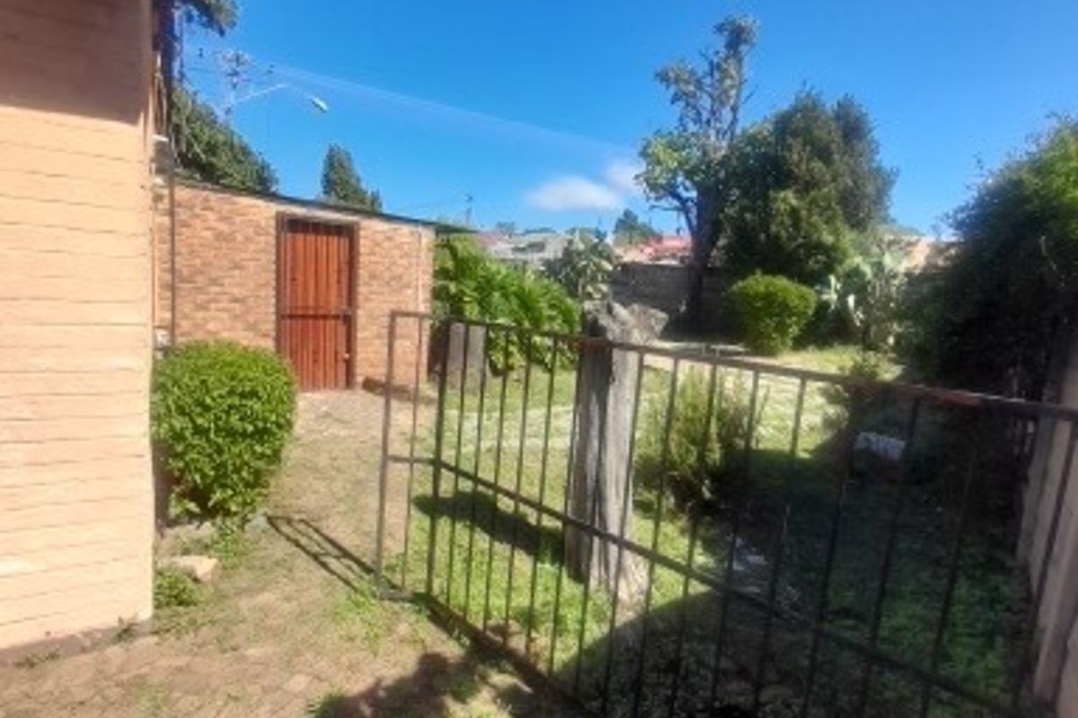 Rear exterior view showing a brick-paved area, a metal gate, and a small brick outbuilding set within a garden space.