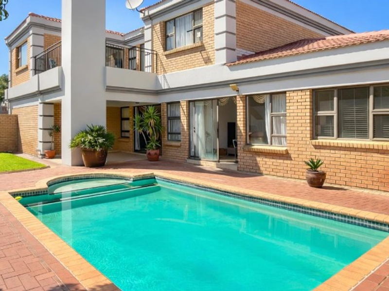 A well-maintained rear exterior featuring a swimming pool, brick paved patio, and a two-story brick residence with a balcony and chimney.