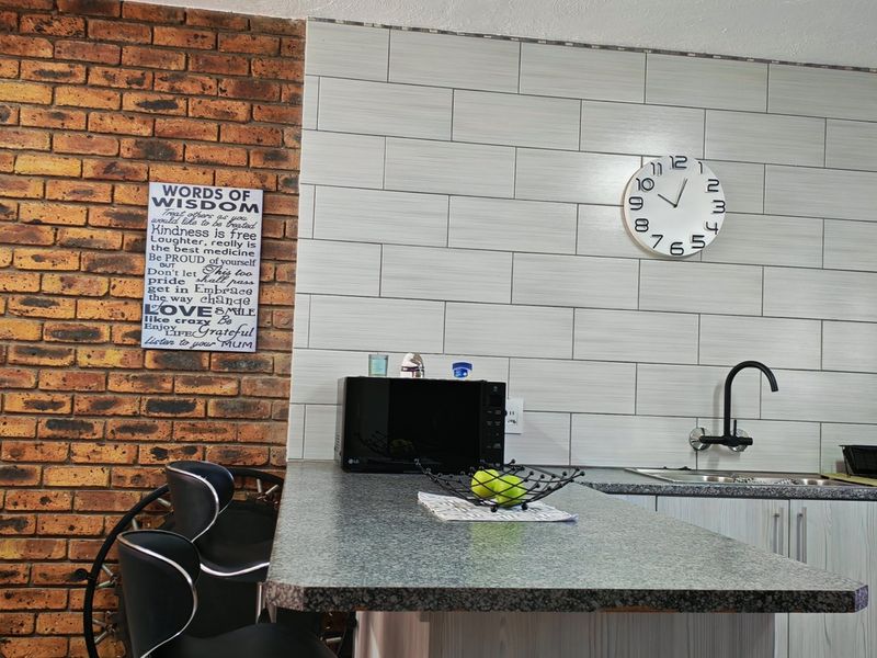A modern kitchen area featuring a striking exposed brick feature wall, light-toned tiled backsplash, granite countertops, and a sleek black mixer tap.
