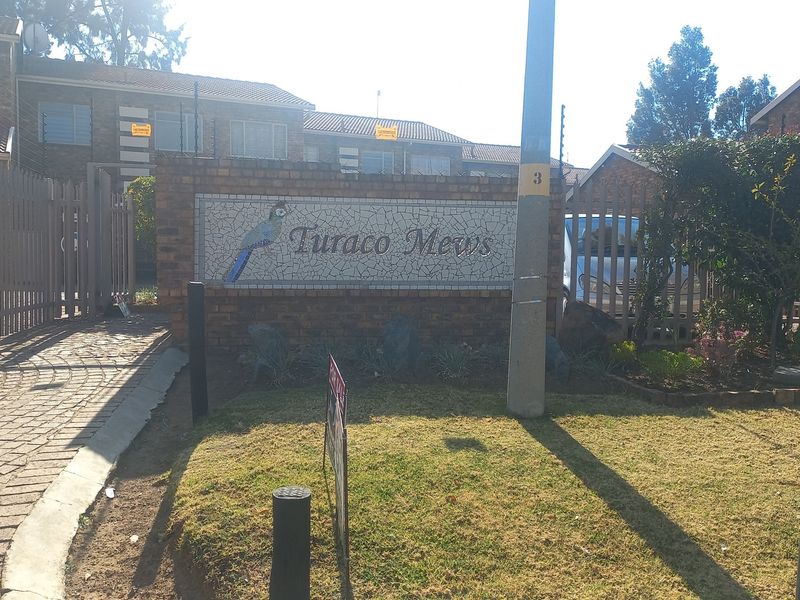 Front entrance view of Turaco Mews townhouse complex featuring a brick boundary wall, electric fencing, and paved driveway access.