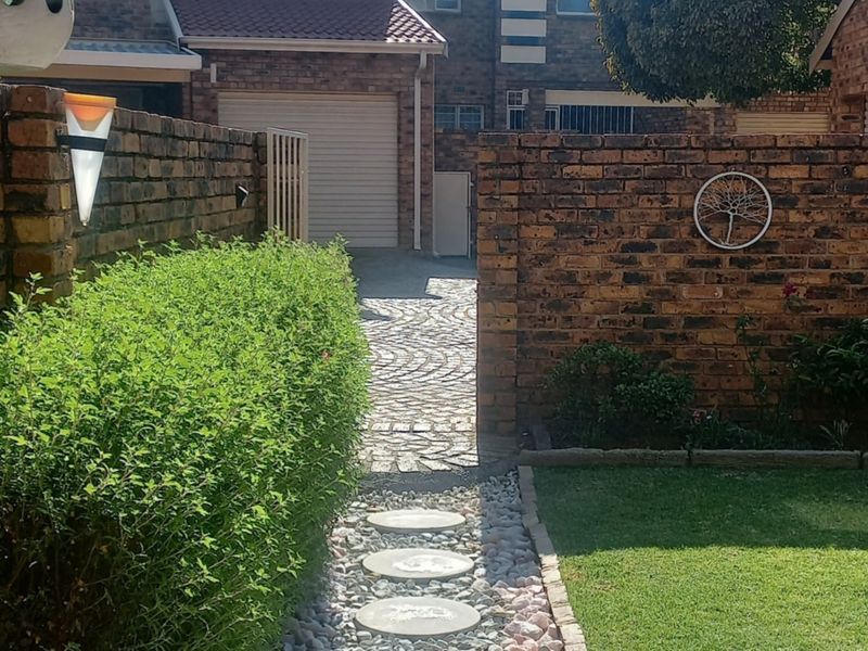 A view of the front exterior of the townhouse, featuring a brick boundary wall, paved driveway, garage access, and a landscaped garden path.