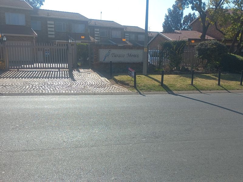 Street view of the Turaco Mews townhouse complex entrance, featuring brick paving, security gates, and established perimeter fencing.