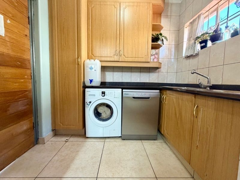 A functional scullery featuring wood-finish cabinetry, dark laminate countertops, a stainless steel sink, and tiled splashbacks with space for laundry appliances.
