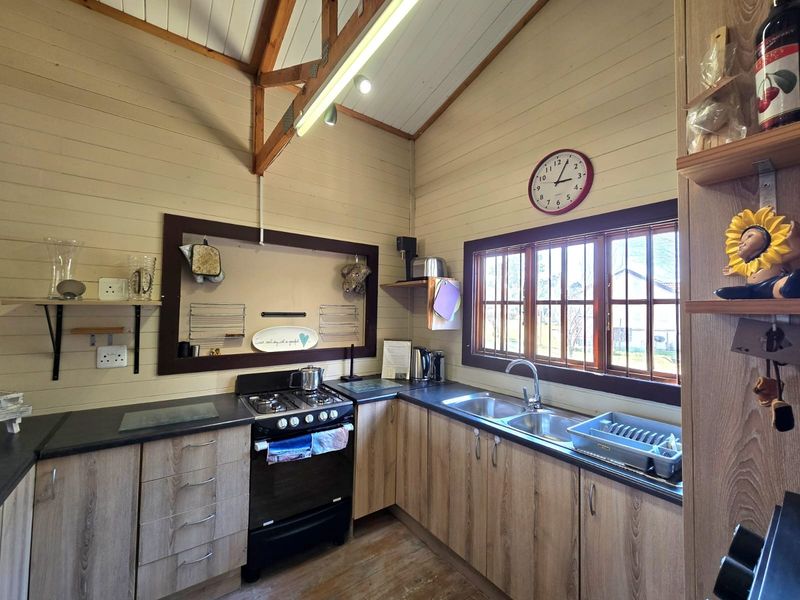 A rustic kitchen featuring timber wall cladding, exposed trusses, laminate cabinetry, a gas stove, and a double basin sink with timber-framed windows.