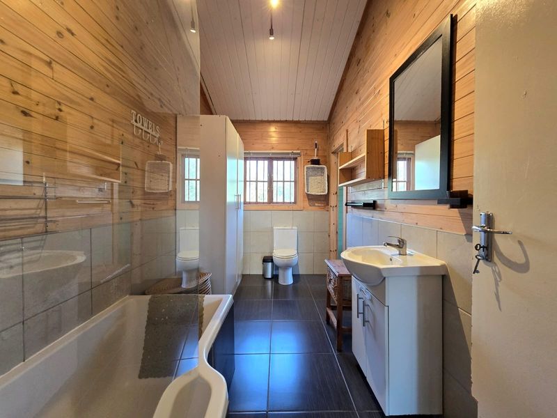 A well-maintained bathroom featuring timber wall cladding, a bathtub with a glass screen, a vanity basin, and a toilet with tiled flooring.