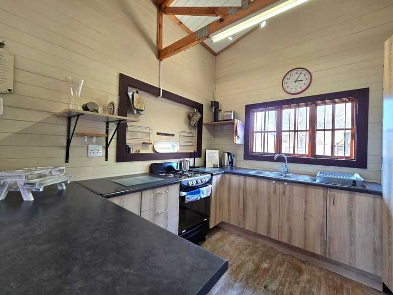 A rustic kitchen featuring timber wall cladding, exposed roof trusses, laminate cabinetry, dark countertops, and a gas stove with a functional layout.