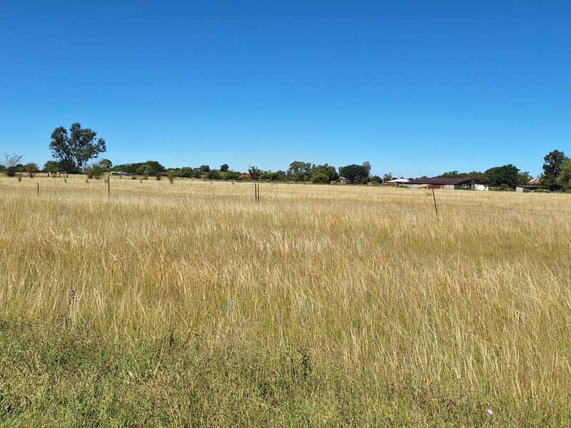 A wide-angle view of a vacant residential plot featuring open grassland and perimeter wire fencing under a clear blue sky.