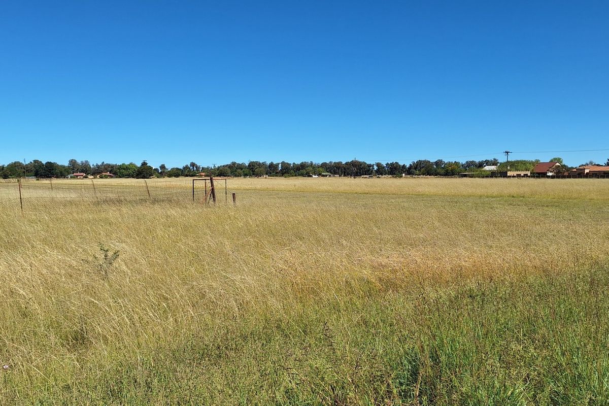 A wide, open vacant residential plot featuring dry grass, perimeter wire fencing, and a metal gate frame.