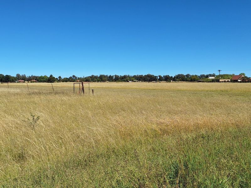 A wide, open vacant residential plot featuring dry grass, perimeter wire fencing, and a metal gate frame.