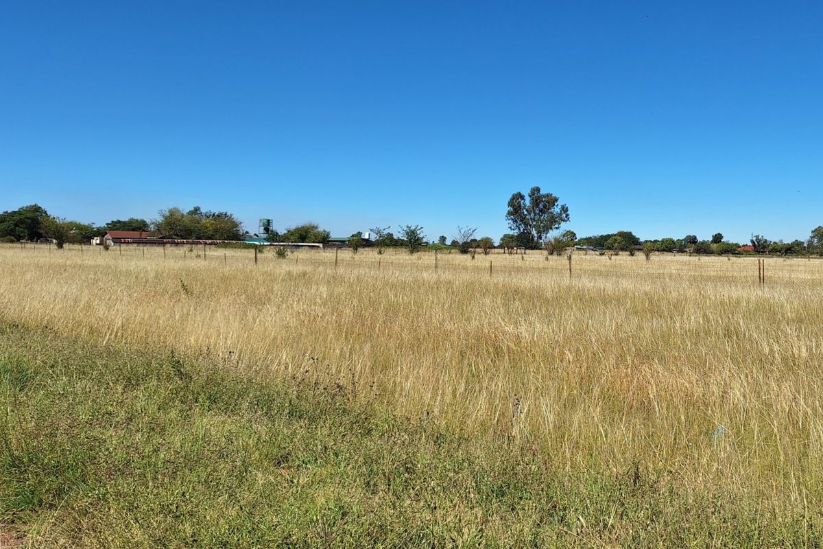 A wide, open vacant residential plot featuring natural grass cover and perimeter wire fencing under a clear blue sky.