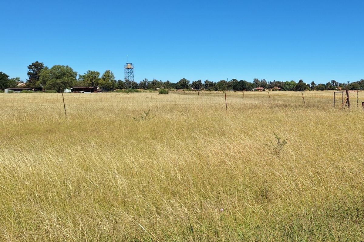 A wide, open residential plot featuring dry grass, perimeter wire fencing, and a prominent water tower structure in the distance.