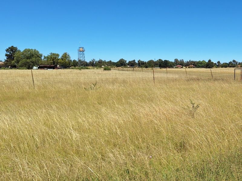 A wide, open residential plot featuring dry grass, perimeter wire fencing, and a prominent water tower structure in the distance.