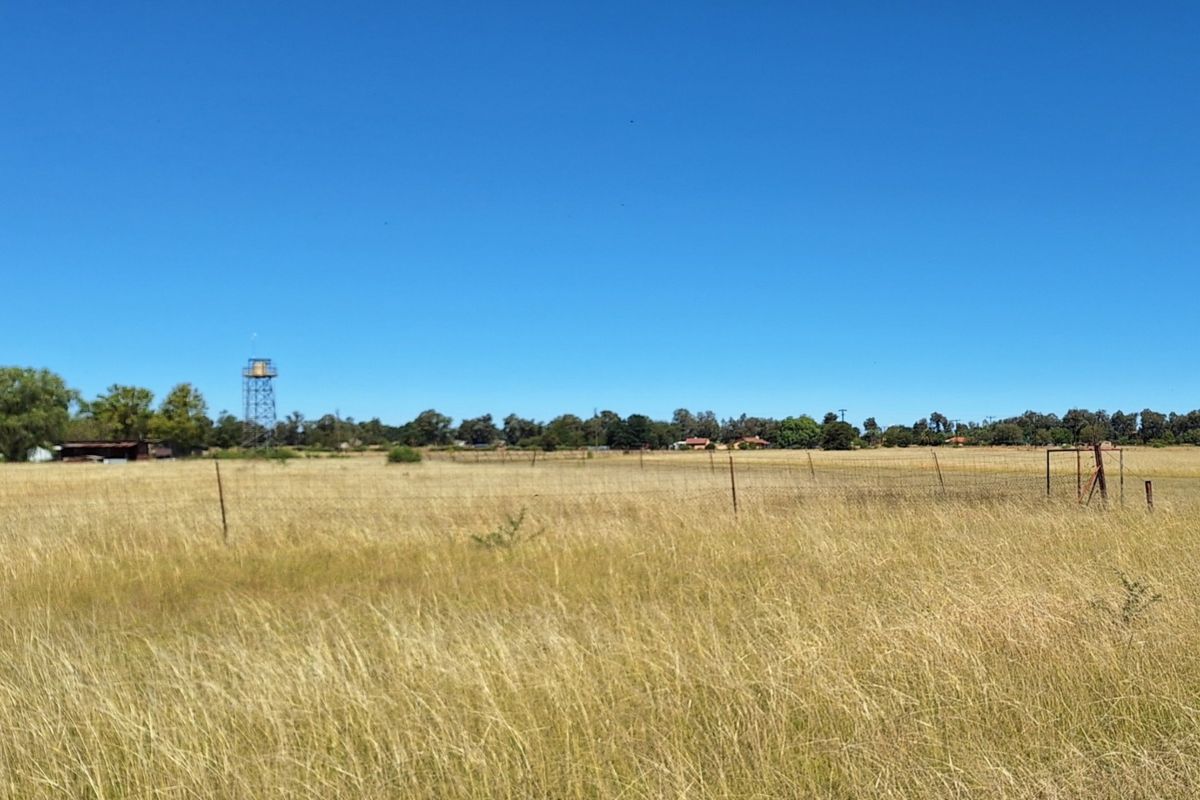 A wide-angle view of a vacant residential plot featuring dry grass, perimeter wire fencing, and a distant water tower structure.