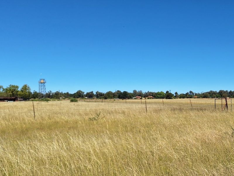 A wide-angle view of a vacant residential plot featuring dry grass, perimeter wire fencing, and a distant water tower structure.