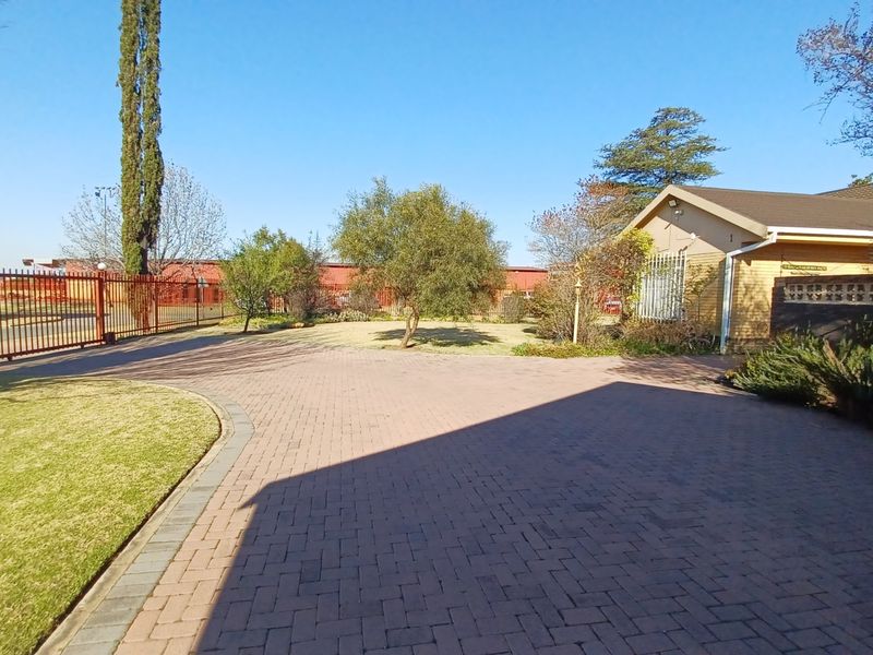 Front exterior view featuring a paved driveway, landscaped garden area, and secure perimeter fencing leading to the main house entrance.