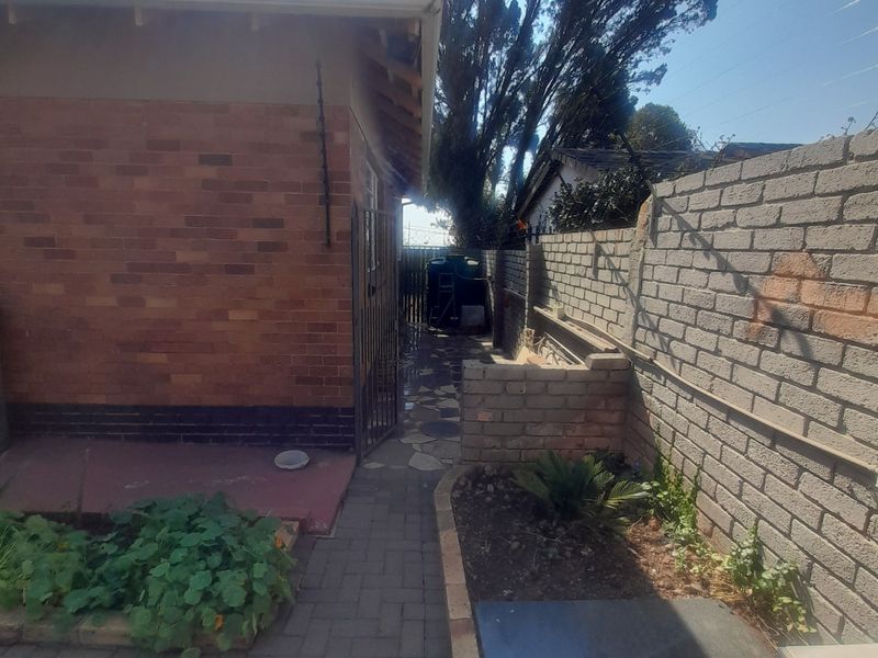 A side exterior view of the property featuring a brick facade, paved walkway, and boundary wall with a security gate.