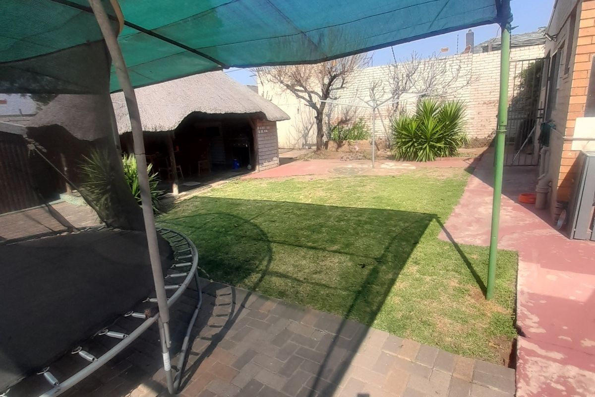 Rear garden area featuring a paved patio, a large shade cloth structure, and a detached thatched lapa in the background.