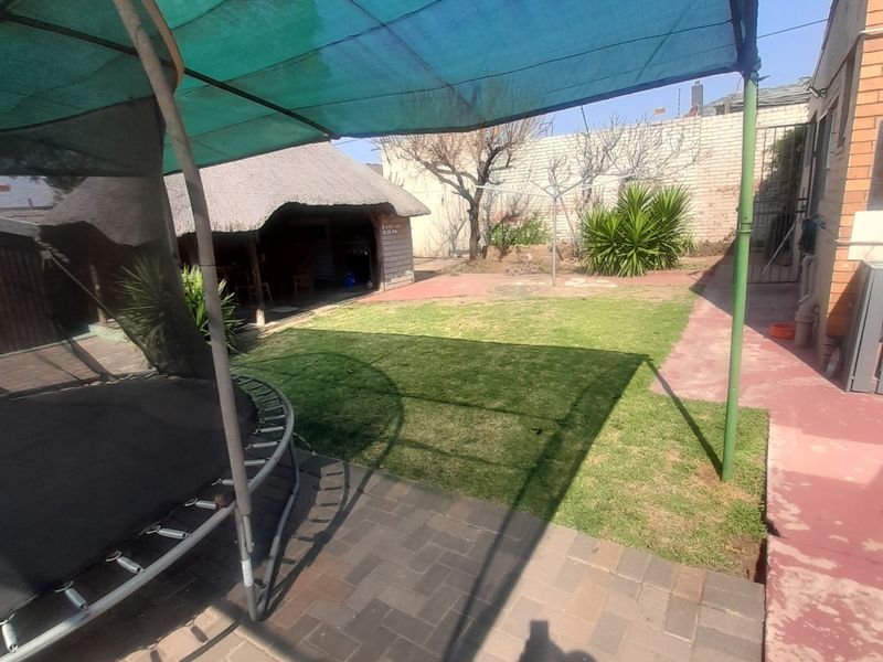 Rear garden area featuring a paved patio, a large shade cloth structure, and a detached thatched lapa in the background.
