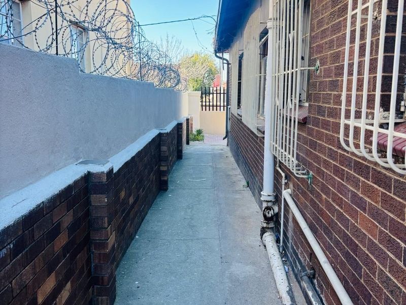 A narrow side passage between two brick buildings featuring security bars on windows, a concrete walkway, and a boundary wall topped with razor wire.