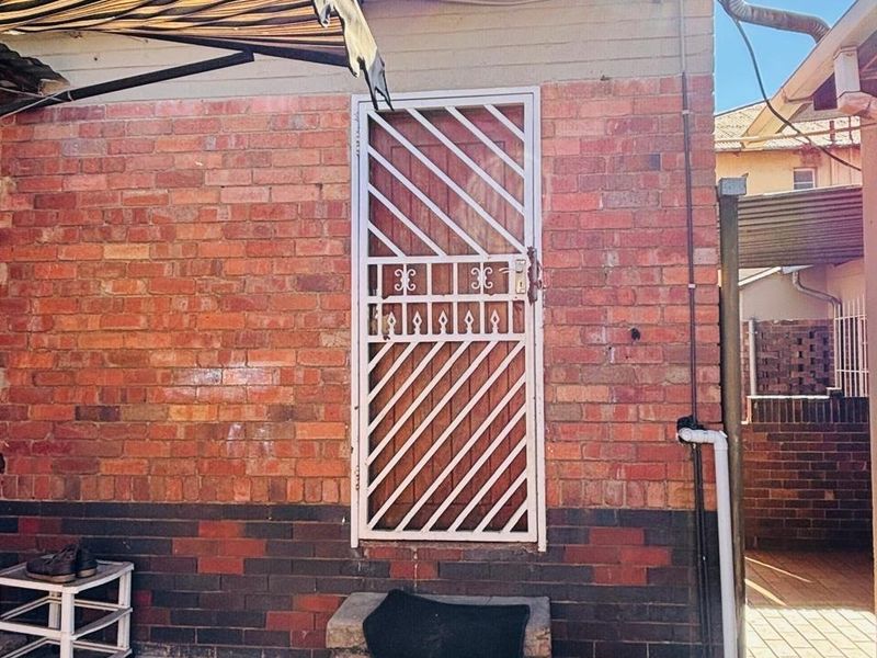 Rear exterior view featuring a brick facade, a security gate entrance, concrete steps, and a retractable awning over a paved courtyard area.