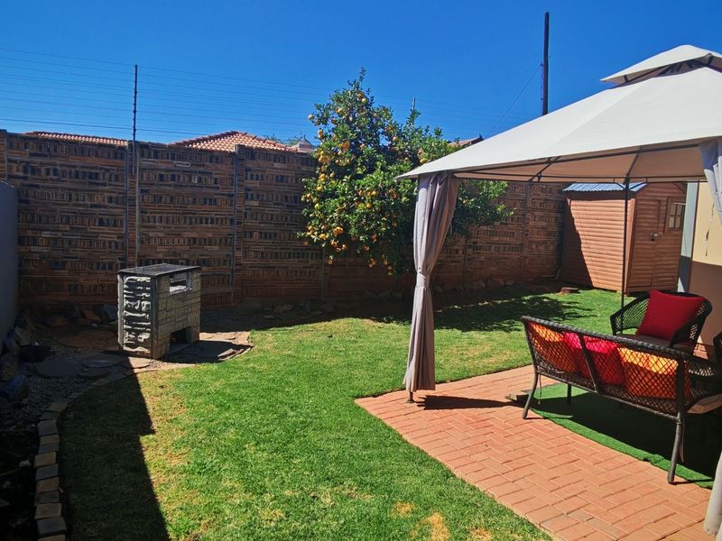 A private rear garden featuring a paved patio area, a built-in braai, and a well-maintained lawn enclosed by brick boundary walls.