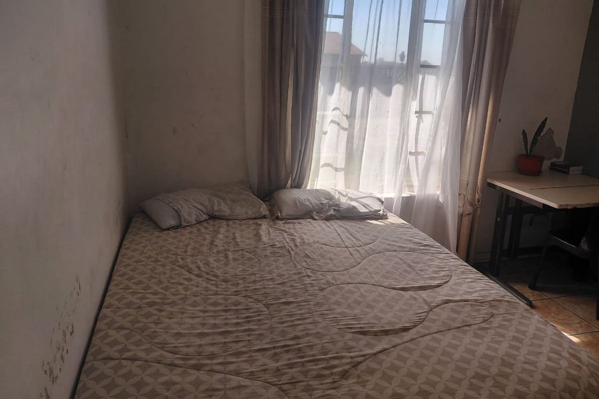 A bedroom featuring tiled flooring and a window, currently showing signs of wall surface damage and peeling paint requiring maintenance.