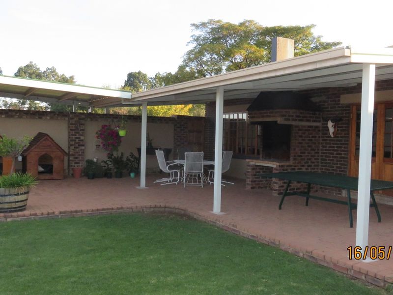 A covered outdoor patio area featuring a built-in brick braai, brick paving, and direct access to the garden through timber framed doors.