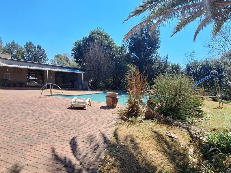 Outdoor pool area featuring a brick-paved patio, covered veranda, and a swimming pool with a slide, set within a garden environment.