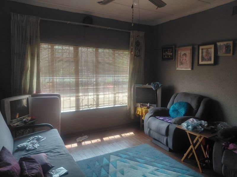A living room featuring timber flooring, a ceiling fan, and large windows with venetian blinds, offering a functional space with potential for modernization.