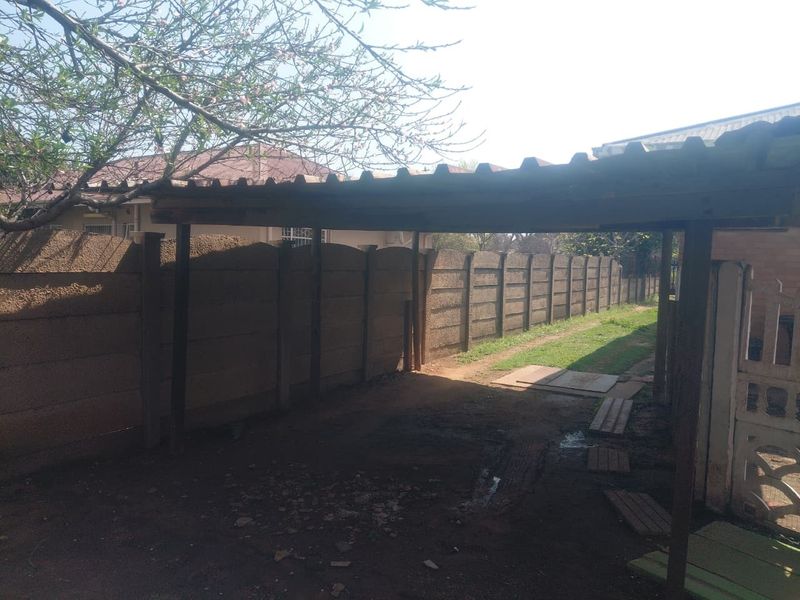 A rear exterior view featuring a basic carport structure and a long precast concrete boundary wall in a residential garden setting.