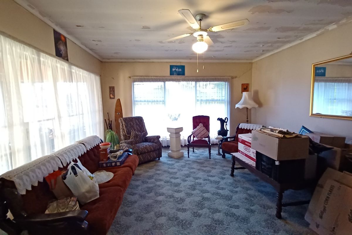 A living room featuring large windows and a ceiling fan, currently showing significant wear to the ceiling paint and flooring.