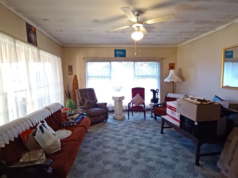 A living room featuring large windows and a ceiling fan, currently showing significant wear to the ceiling paint and flooring.