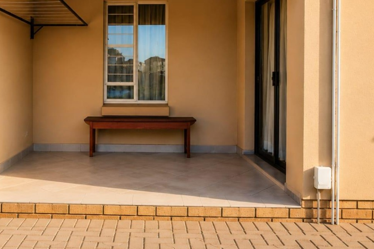 A clean, covered patio area featuring tiled flooring, a sliding glass door entrance, and a wall-mounted drying rack, providing a functional outdoor space.
