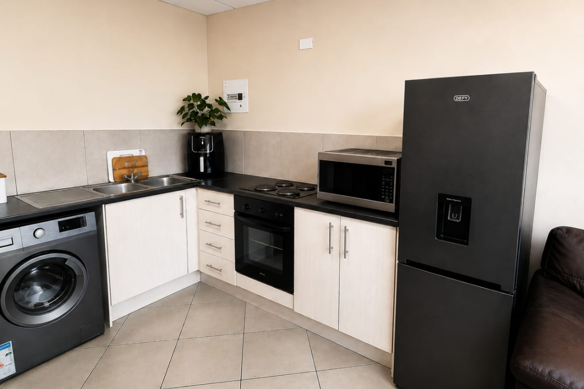 A compact kitchen featuring light-toned cabinetry, dark countertops, a built-in oven and hob, and tiled flooring with integrated laundry and refrigeration space.