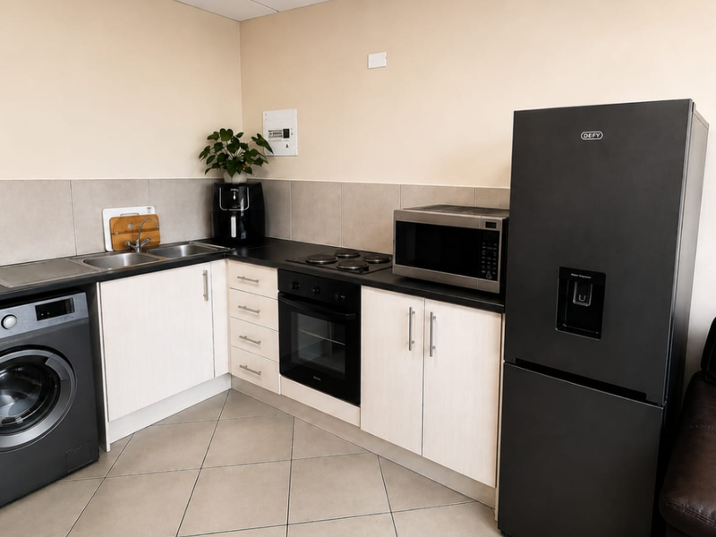 A compact kitchen featuring light-toned cabinetry, dark countertops, a built-in oven and hob, and tiled flooring with integrated laundry and refrigeration space.