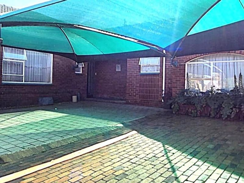 A paved outdoor patio area featuring a large green shade sail, brick exterior walls, and aluminium window frames.