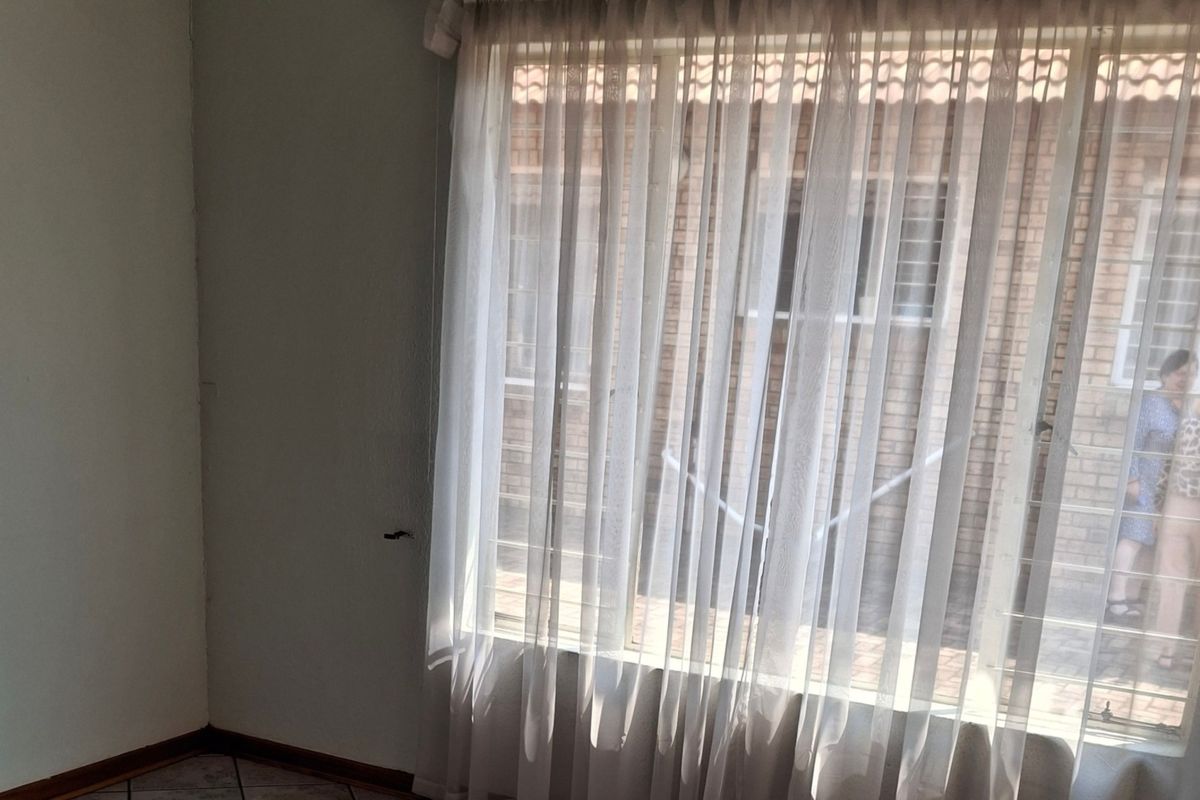 A bright, empty living area featuring tiled flooring and a large window with security bars, offering a view of the exterior brick facade.