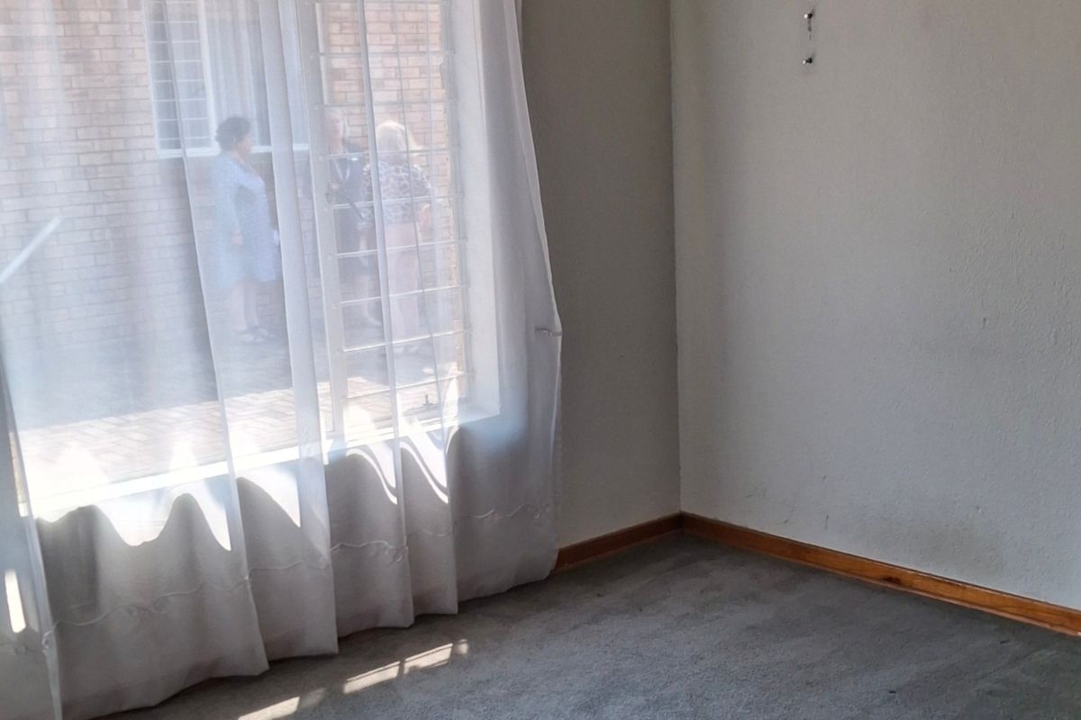 A vacant bedroom featuring neutral walls, carpeted flooring, timber skirting boards, and a large window with an external security gate.