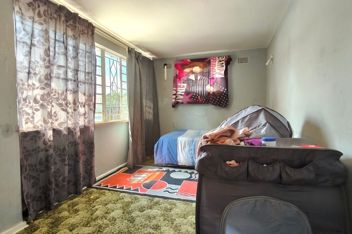 A bedroom featuring a window with security bars and a ventilation grille, currently showing signs of wear on the walls and flooring.