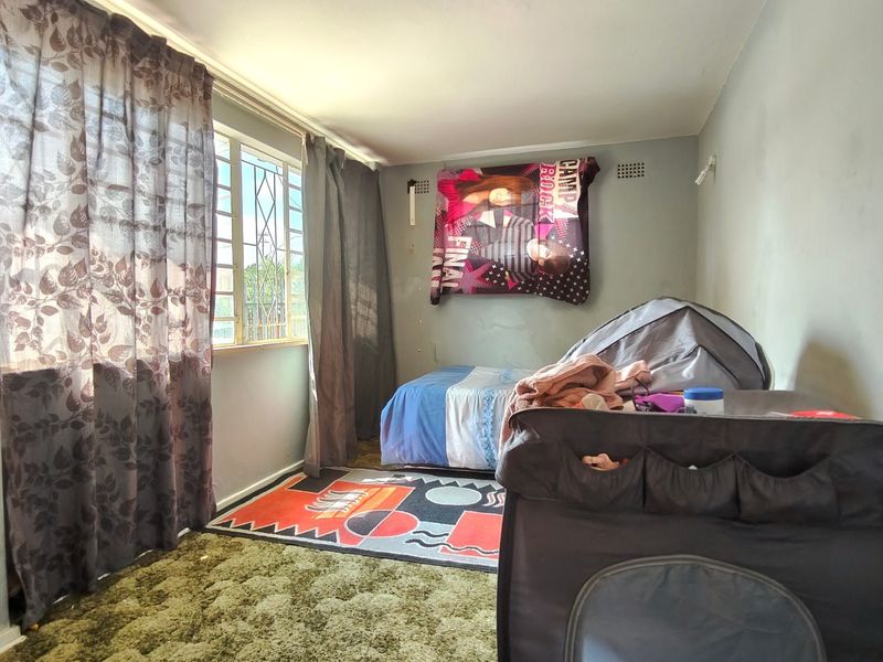 A bedroom featuring a window with security bars and a ventilation grille, currently showing signs of wear on the walls and flooring.
