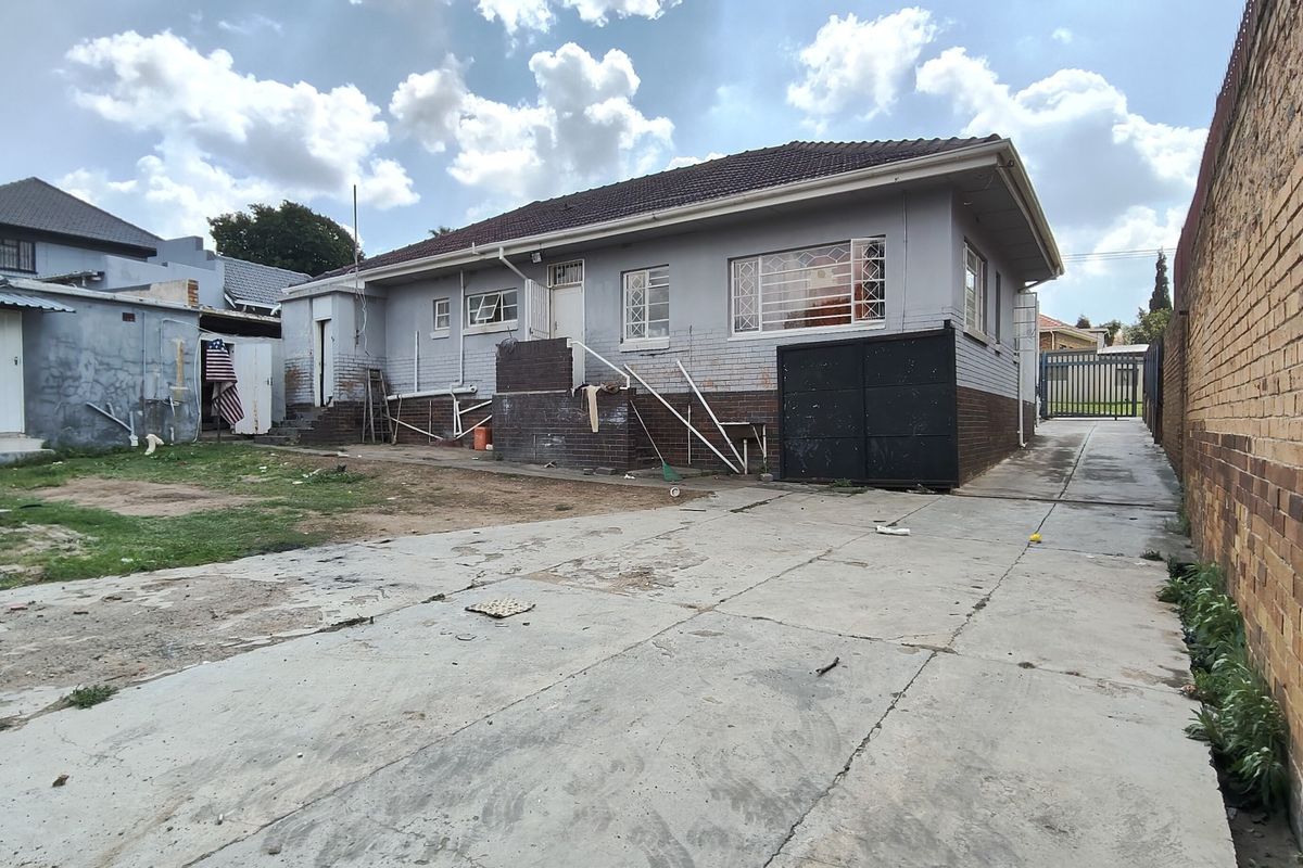 Rear view of the property featuring a concrete driveway, brick boundary wall, and security gate, showing signs of wear and maintenance requirements.
