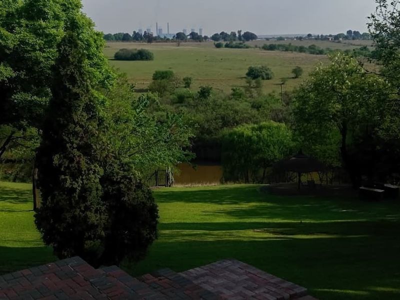 A scenic view overlooking a lush green garden with mature trees, a small pond, and distant city skyline views.