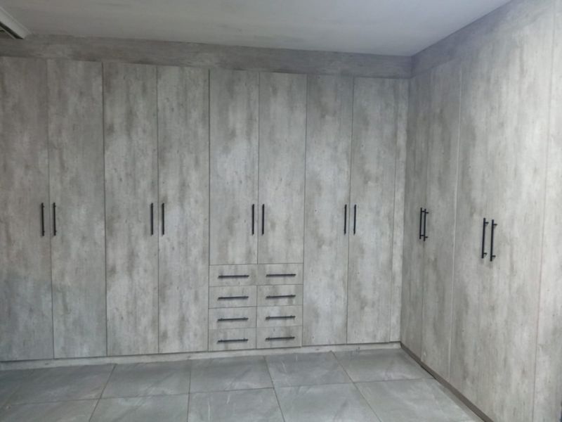 A bedroom featuring extensive floor-to-ceiling built-in cupboards with a modern grey concrete-look finish and dark hardware, complemented by large format floor tiles.