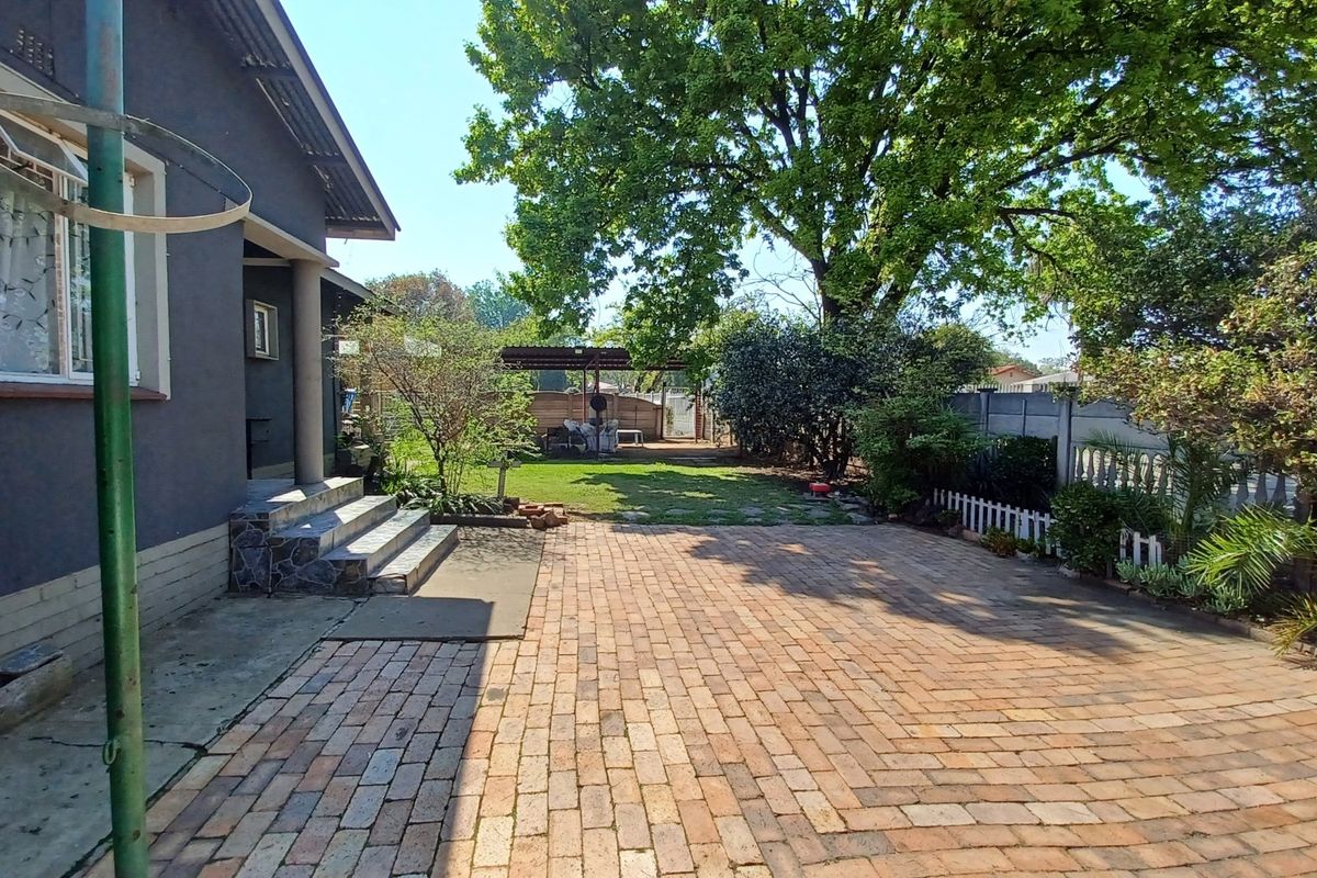A spacious rear garden featuring a brick-paved patio area, concrete steps leading to the house, established lawn, and mature trees.