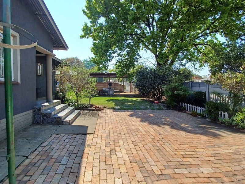 A spacious rear garden featuring a brick-paved patio area, concrete steps leading to the house, established lawn, and mature trees.