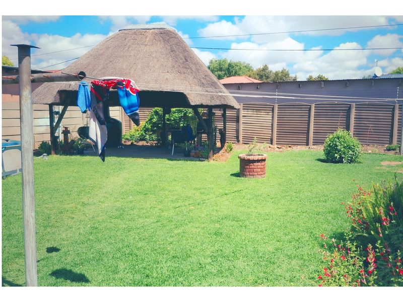 A spacious backyard featuring a traditional thatch-roof lapa, a well-maintained lawn, and perimeter brick walling.