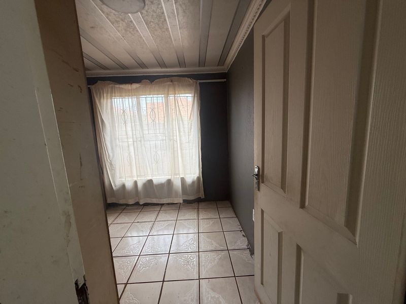 An empty bedroom featuring tiled flooring, a window with security bars, and decorative ceiling cornicing, ready for renovation or staging.