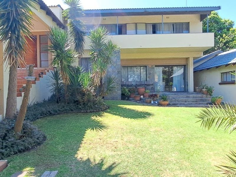 Spacious rear exterior view featuring a landscaped garden, tiled patio area, stone-clad accents, and a multi-level house structure with a balcony.