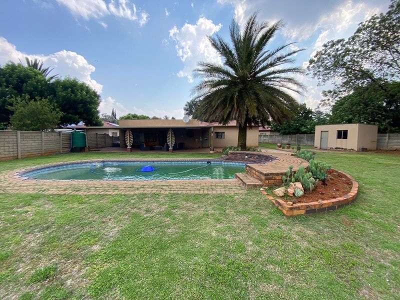 Spacious backyard featuring a swimming pool with brick surround, raised garden bed, covered patio, and a detached outbuilding set within a large lawn area.